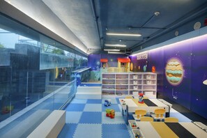 Zona infantil con suelo azul, mesas, sillas y una estantería.