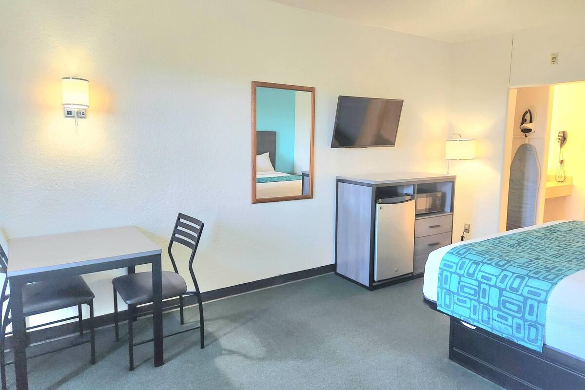 Una habitación de hotel con cama, escritorio con silla, televisión, espejo y armario.