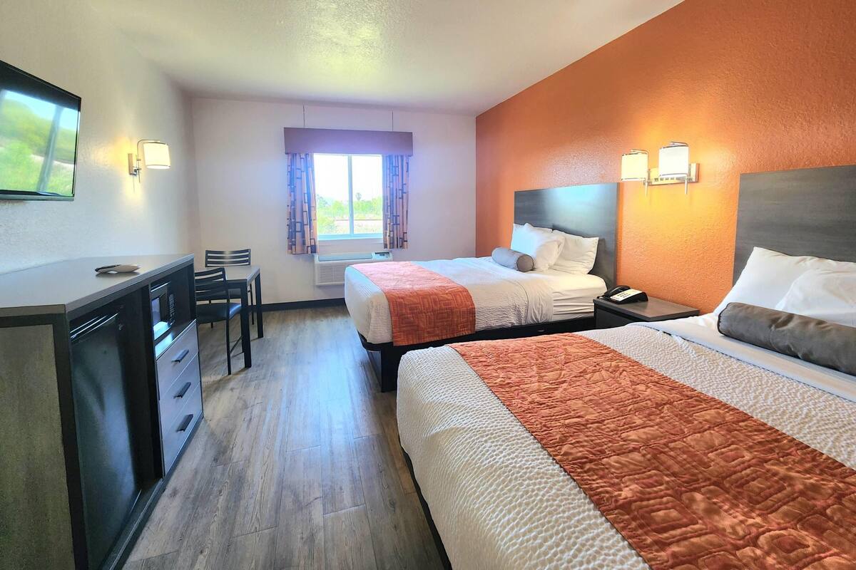 Habitación de hotel con dos camas, un escritorio, sillas, una ventana con cortinas e iluminación en la pared.