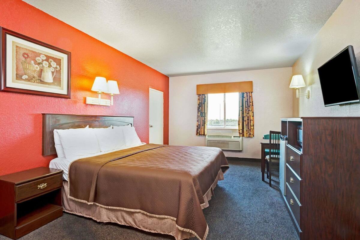 Habitación de hotel con cama, mesita de noche, cómoda, televisión, ventana con cortinas y un cuadro en la pared.