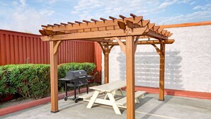 Una pérgola de madera con una mesa de picnic debajo, una parrilla y una pared roja al fondo.