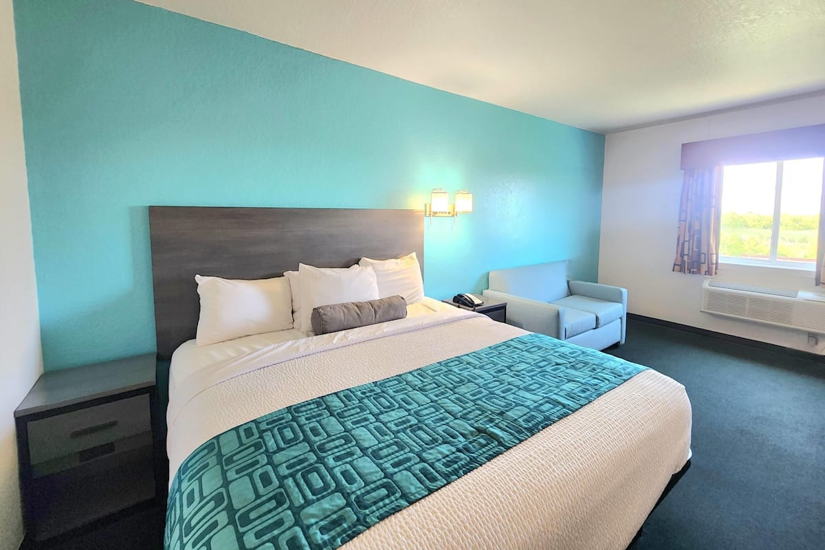 Habitación de hotel con cama, mesita de noche, una silla, ventana con cortinas y lámpara fijada a la pared.