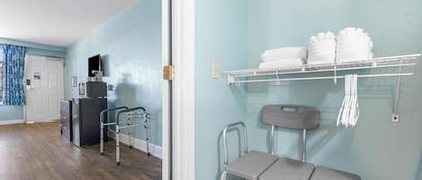 A room with a wooden floor, a white door, a small kitchenette, a TV, a shelf with towels, and a wheelchair.