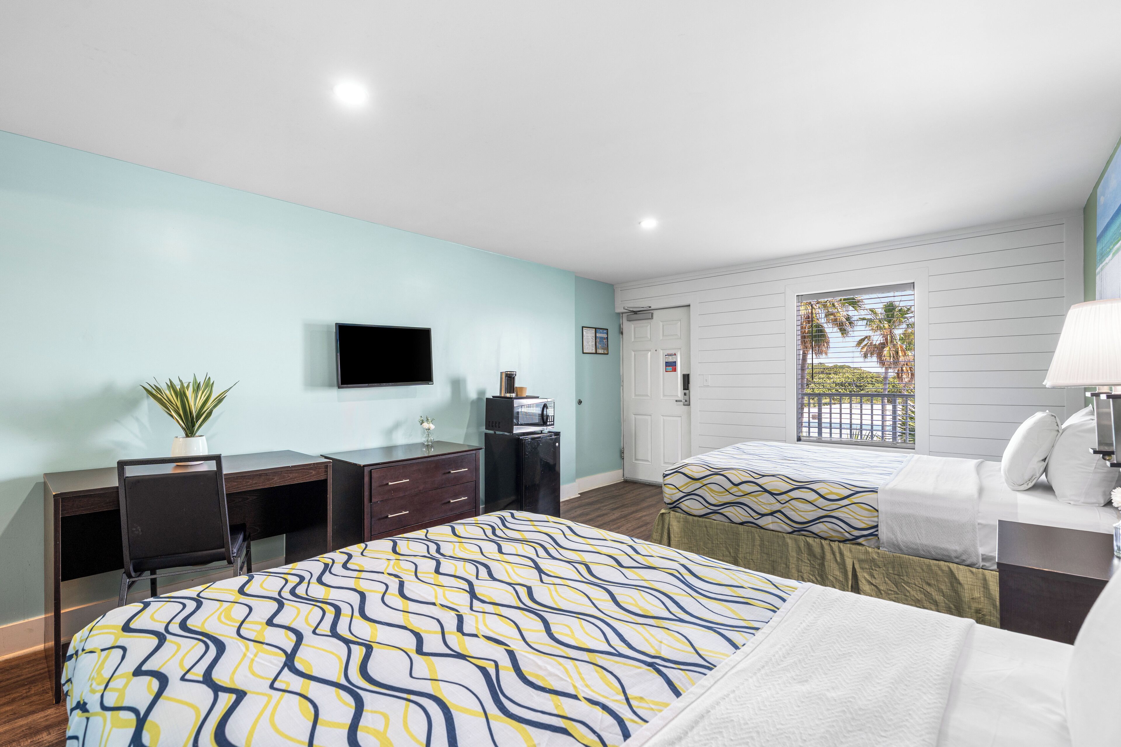 A hotel room with two beds, a desk, a TV, and a window with a view of palm trees.