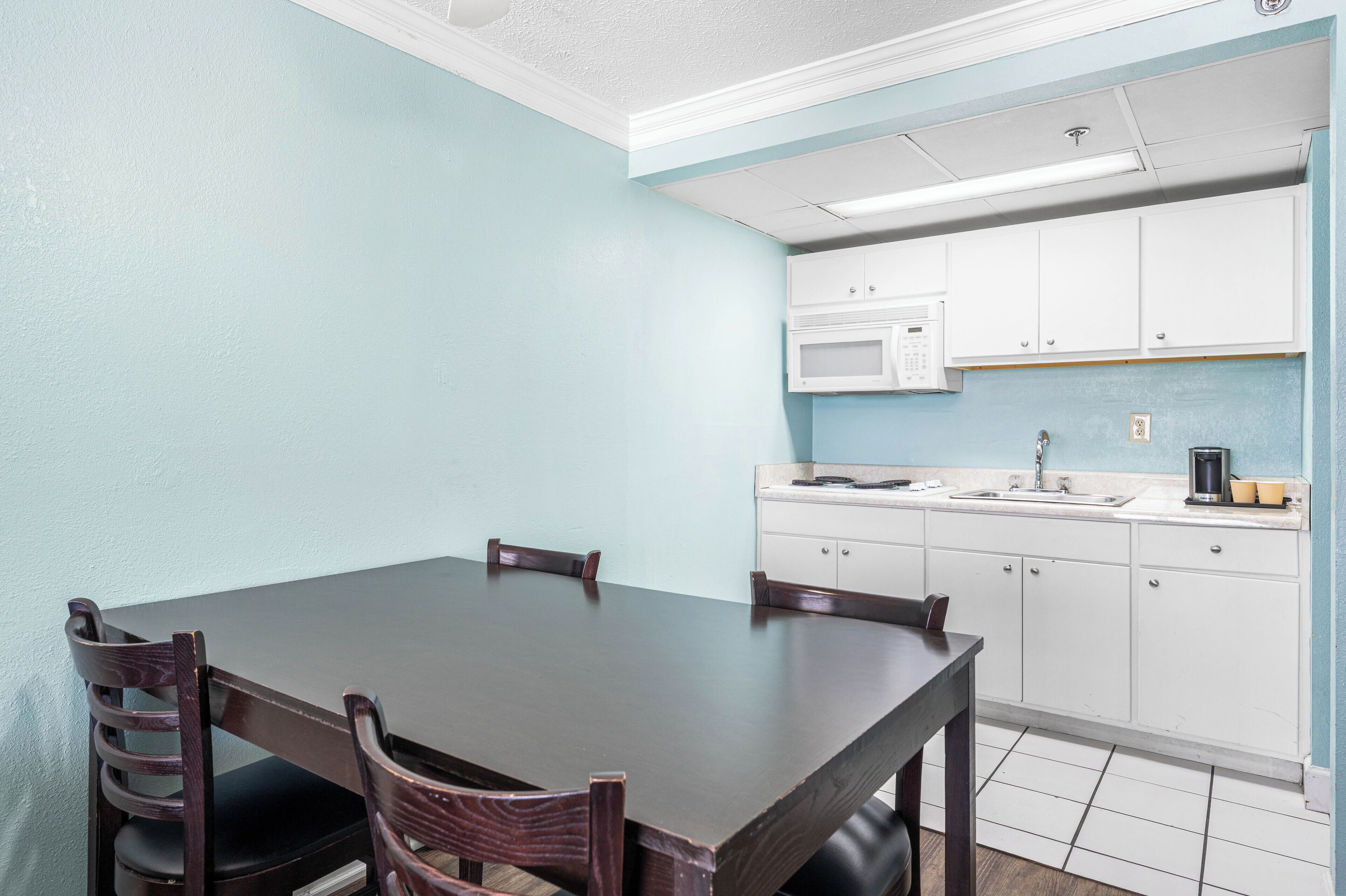 A dining area with a dark wooden table and four chairs, a kitchen with white cabinets and a microwave, and a light blue wall.