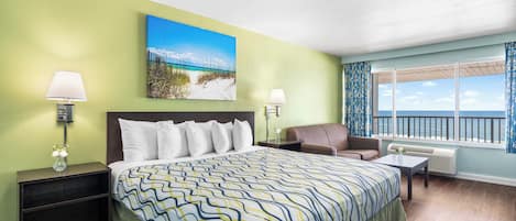A hotel room with a bed, a chair, a nightstand, a lamp, a painting, and a window with a view of the ocean.