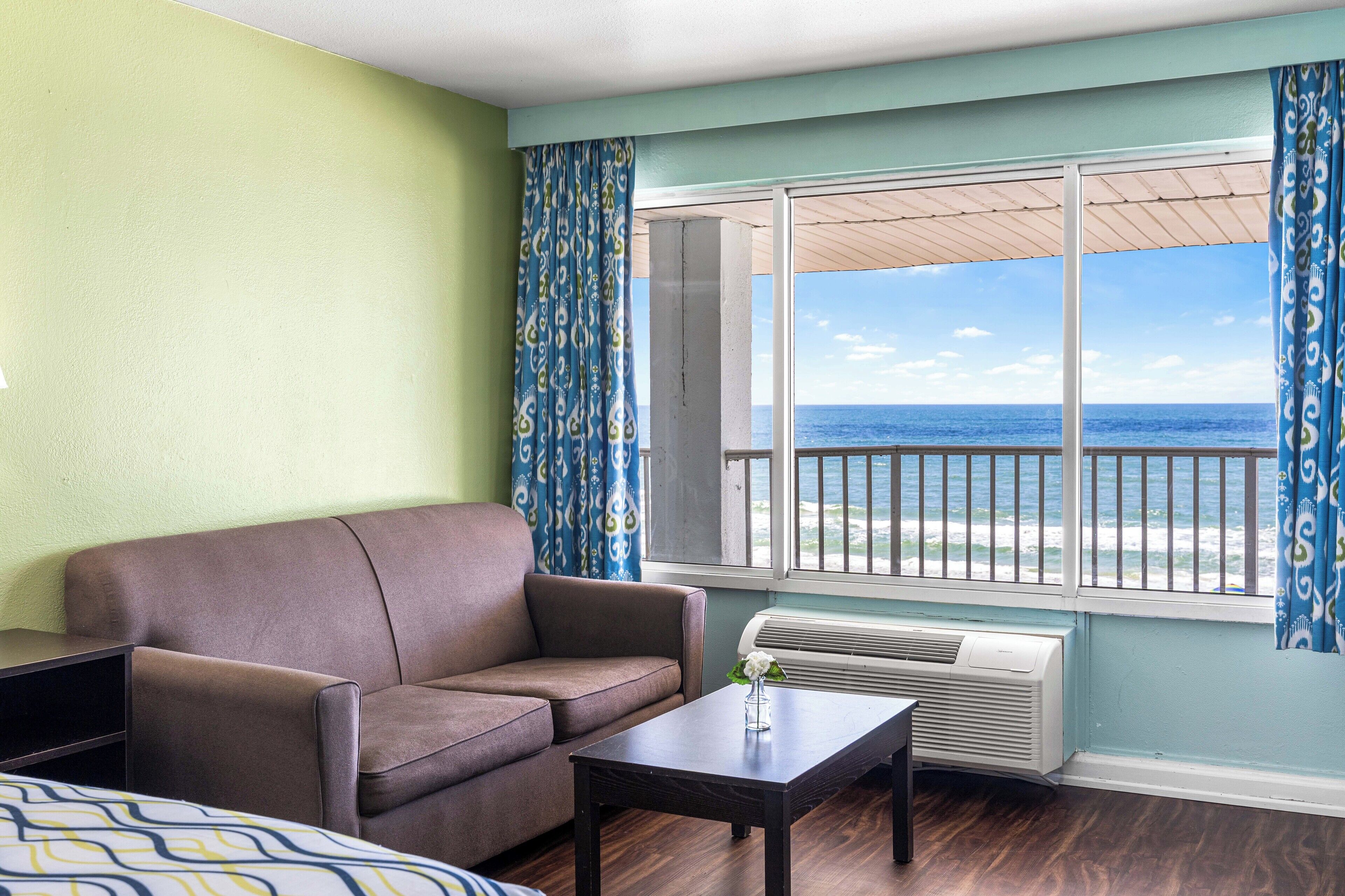 A room with a brown sofa, a small table with a vase, and a large window with a view of the ocean.