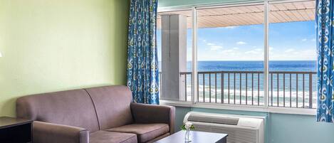 A room with a brown sofa, a small table with a vase, and a large window with a view of the ocean.