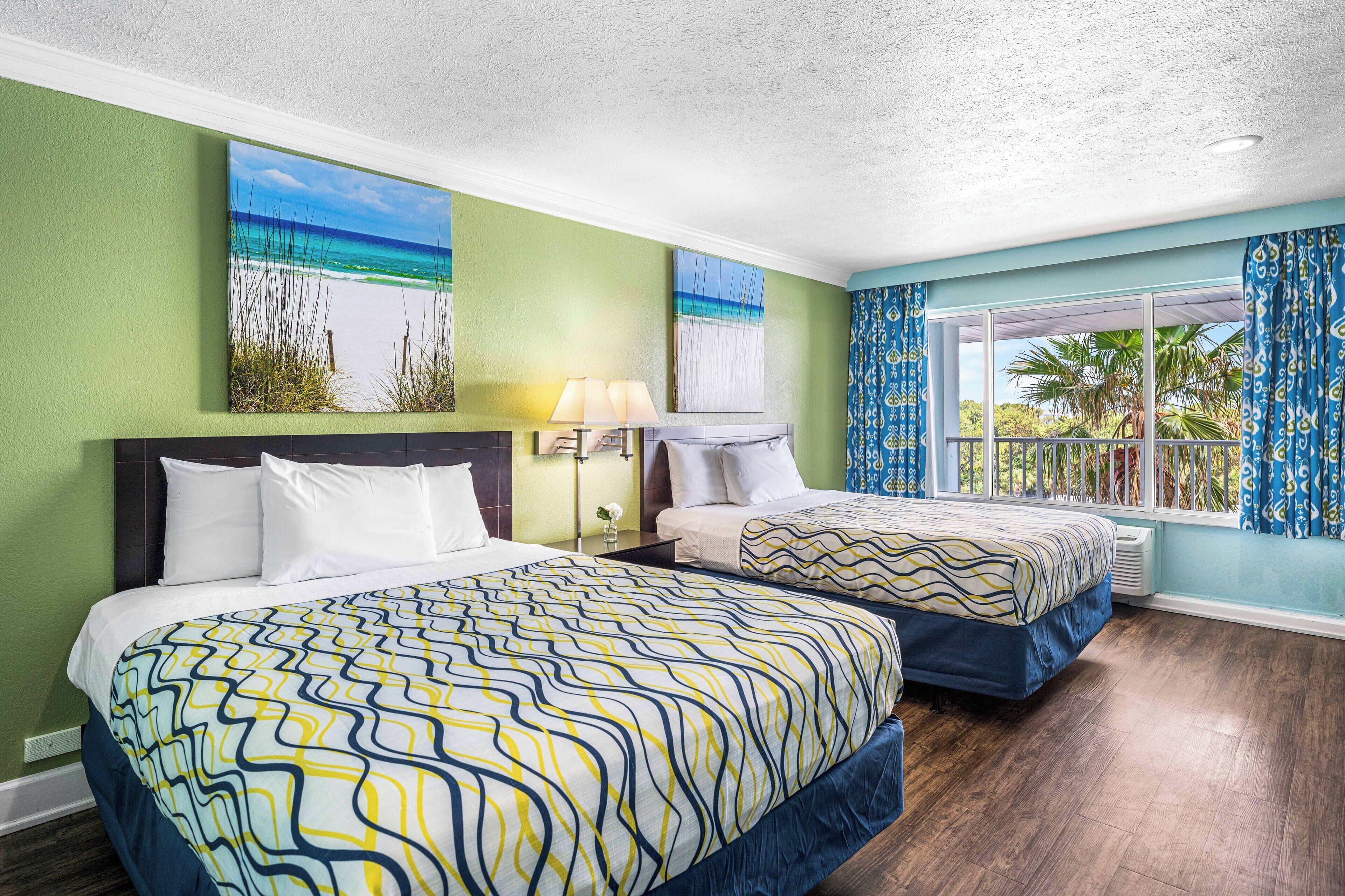 Habitación de hotel con dos camas, paredes verdes, un cuadro con motivos playeros, una ventana con cortinas y vistas a palmeras.