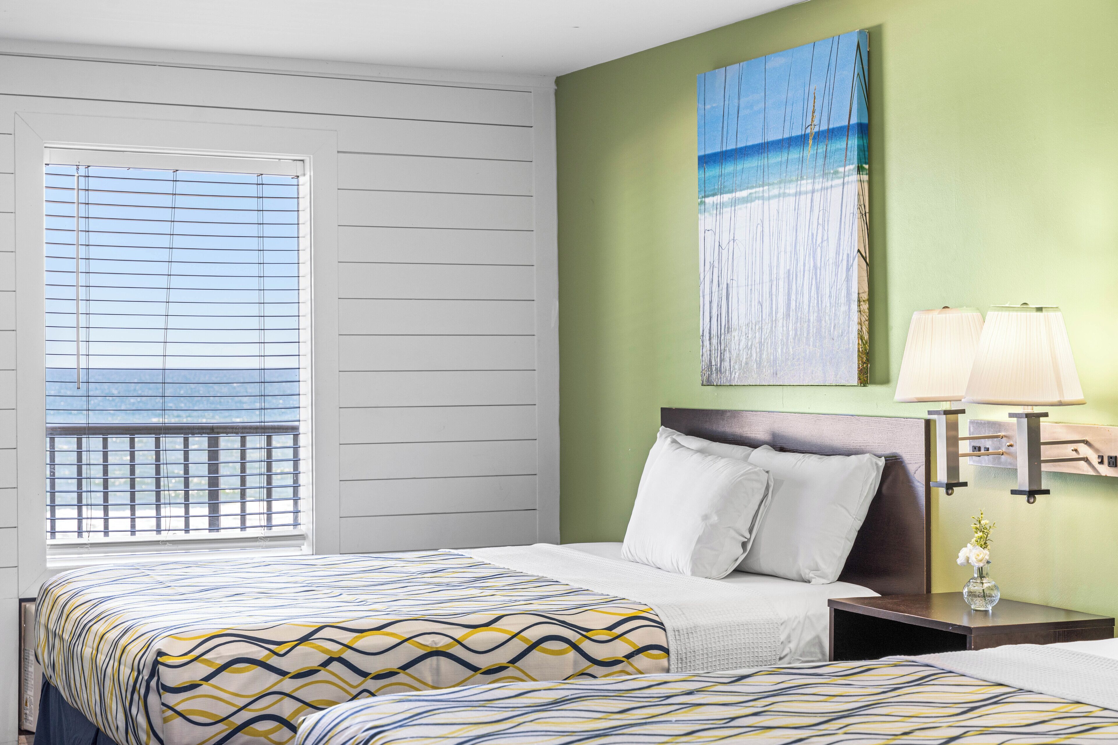 A hotel room with a bed, a nightstand with a lamp, a painting on the wall, and a window with blinds.