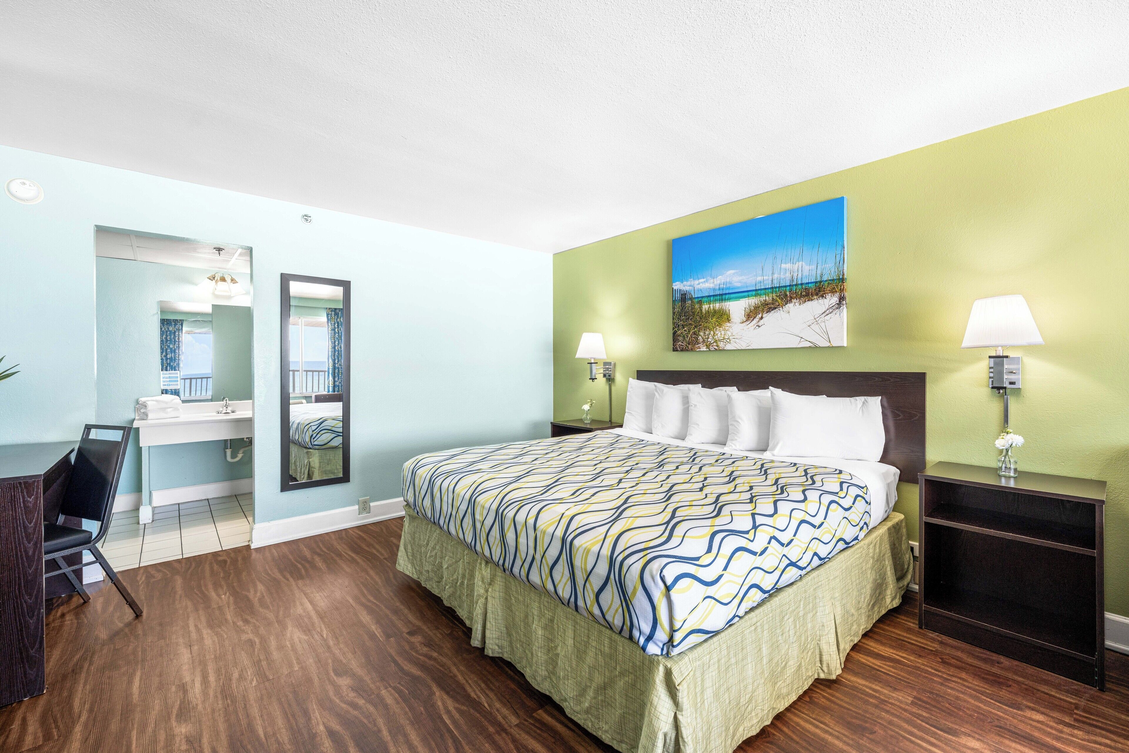 A hotel room with a bed, a desk, a chair, a nightstand, and a painting on the wall.