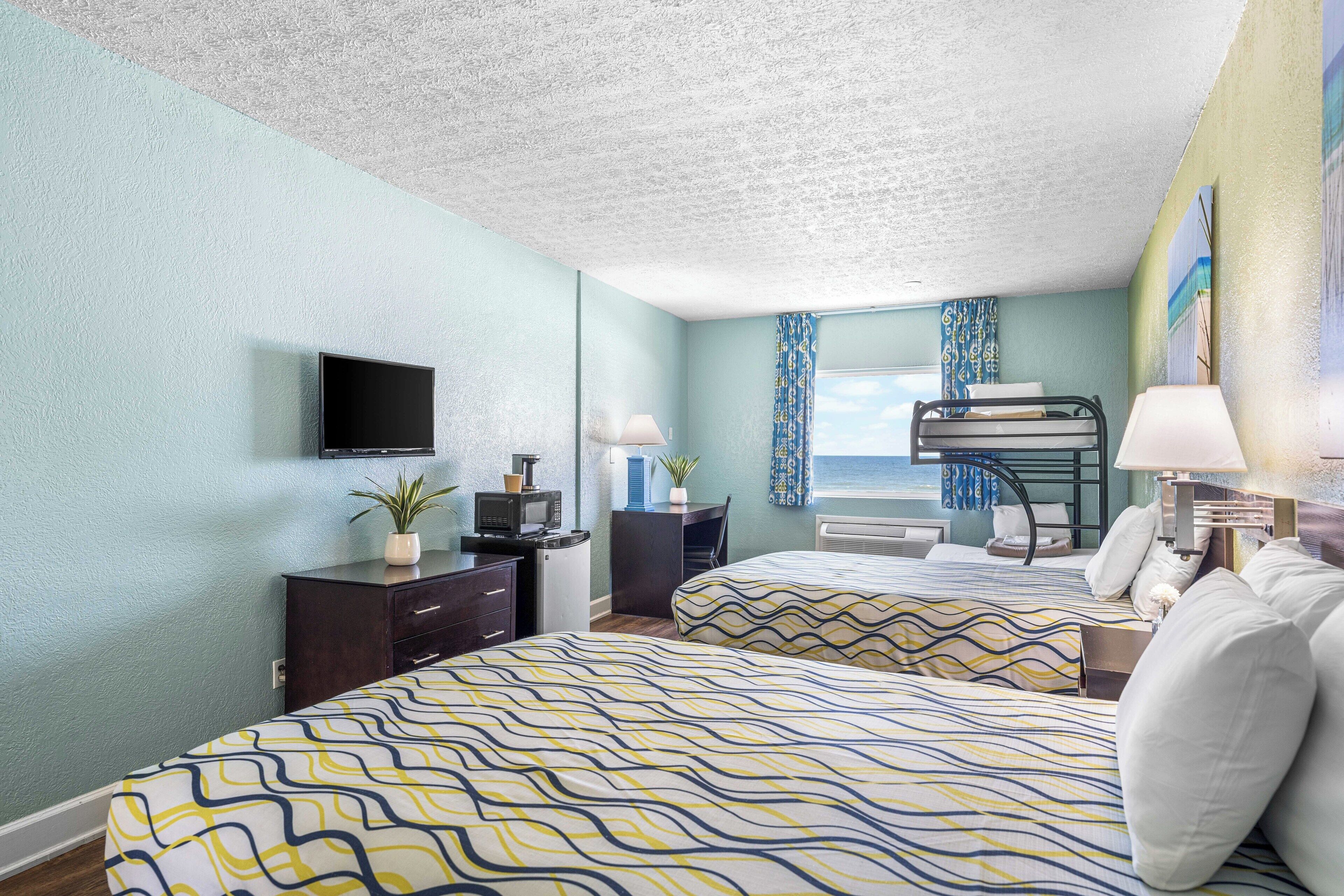 A hotel room with two beds, a desk, a television, and a view of the sea.
