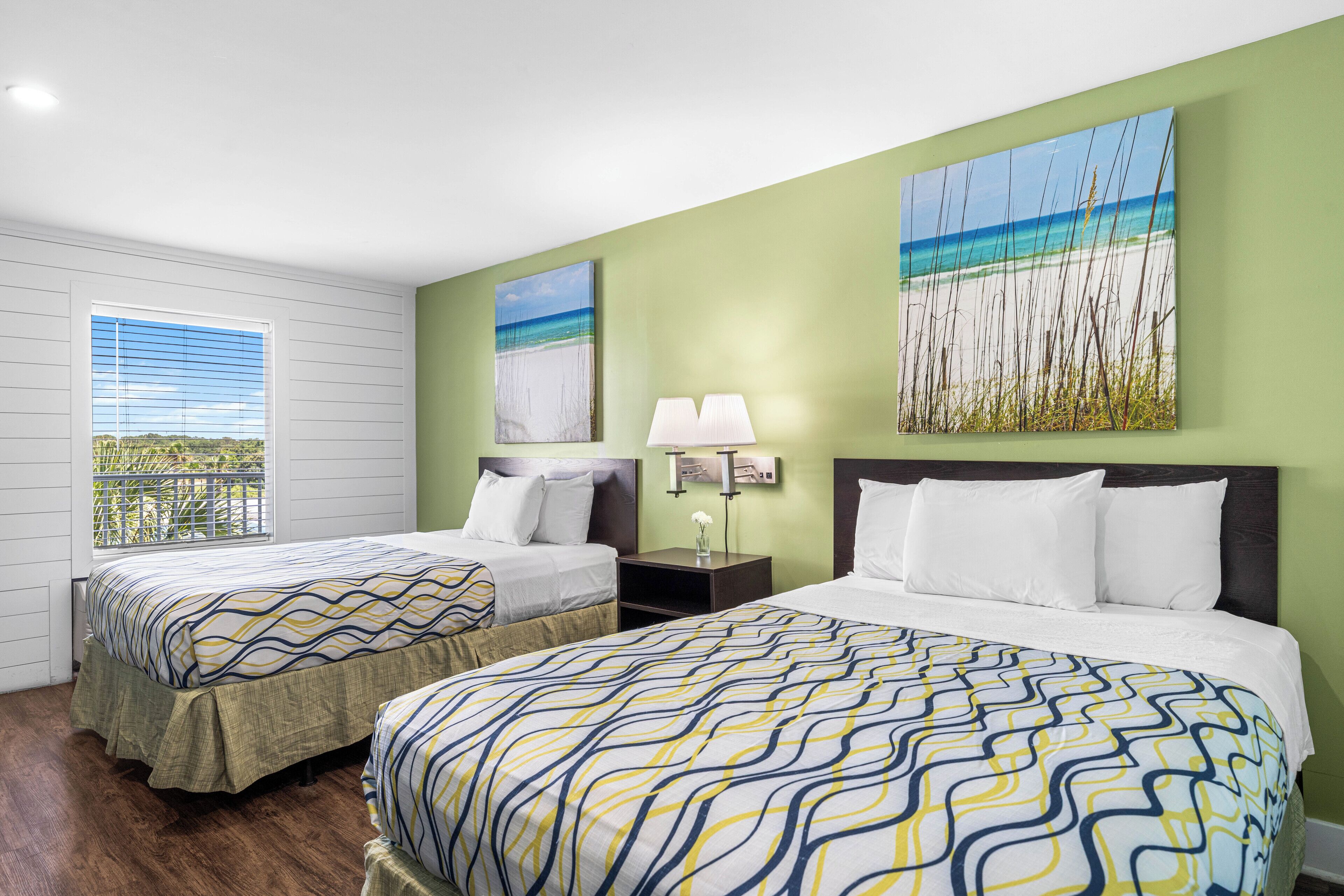 A hotel room with two beds, a window with blinds, a nightstand with a lamp, and artwork on the walls.