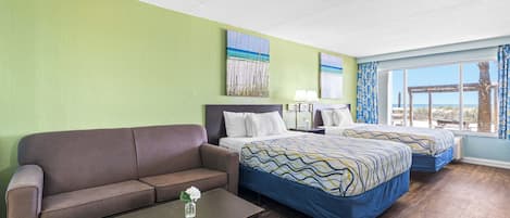 A hotel room with a green wall, two beds with patterned bedding, a brown sofa, a wooden coffee table, and a view of the outdoors through large windows.