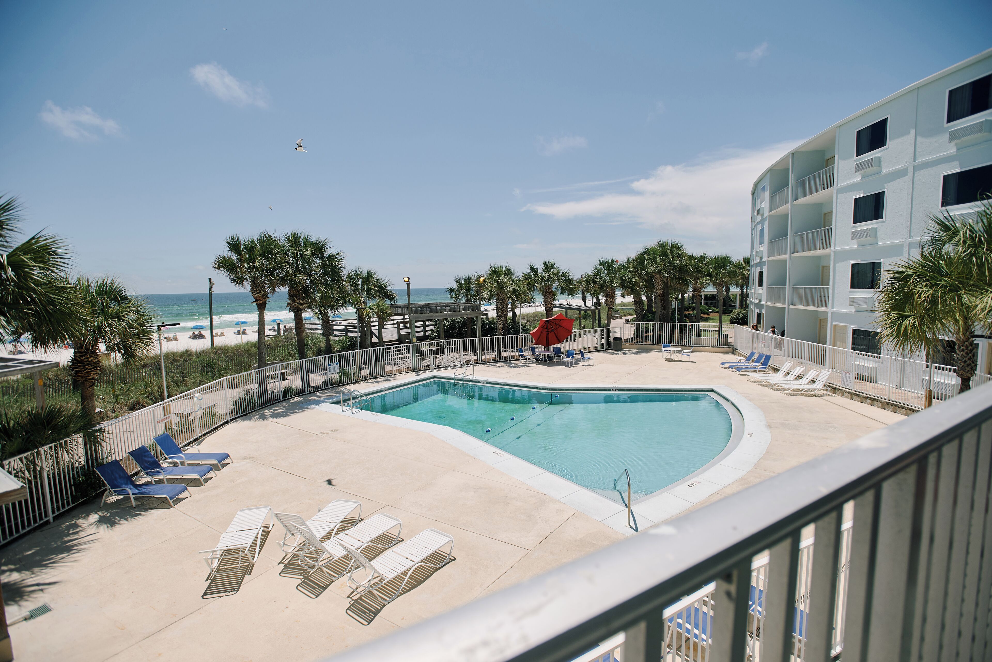 A pool area with lounge chairs and a red umbrella, surrounded by palm trees and a building with balconies.