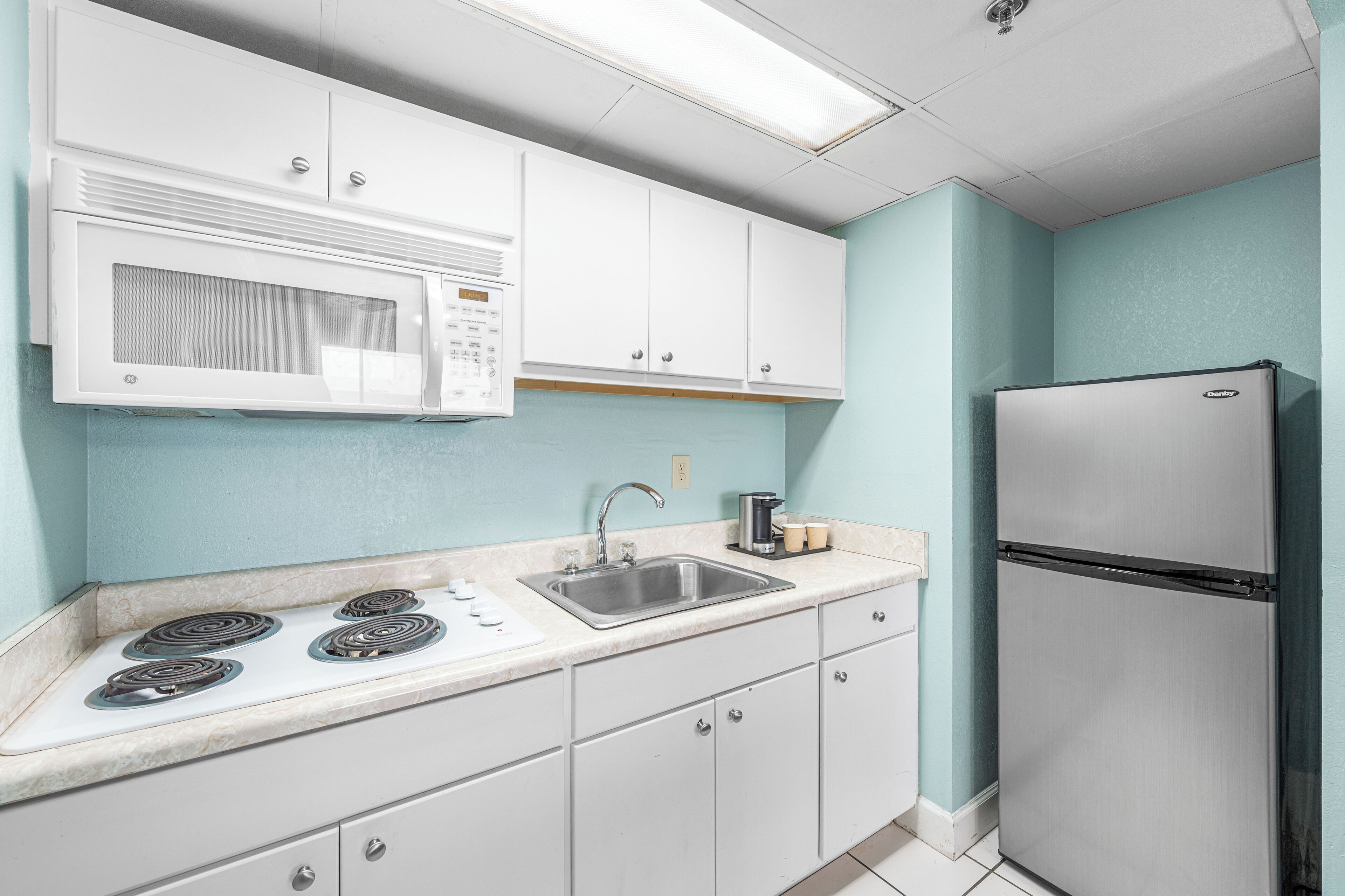 Una cocina con gabinetes blancos, un refrigerador de acero inoxidable, un microondas, un lavamanos y una estufa.