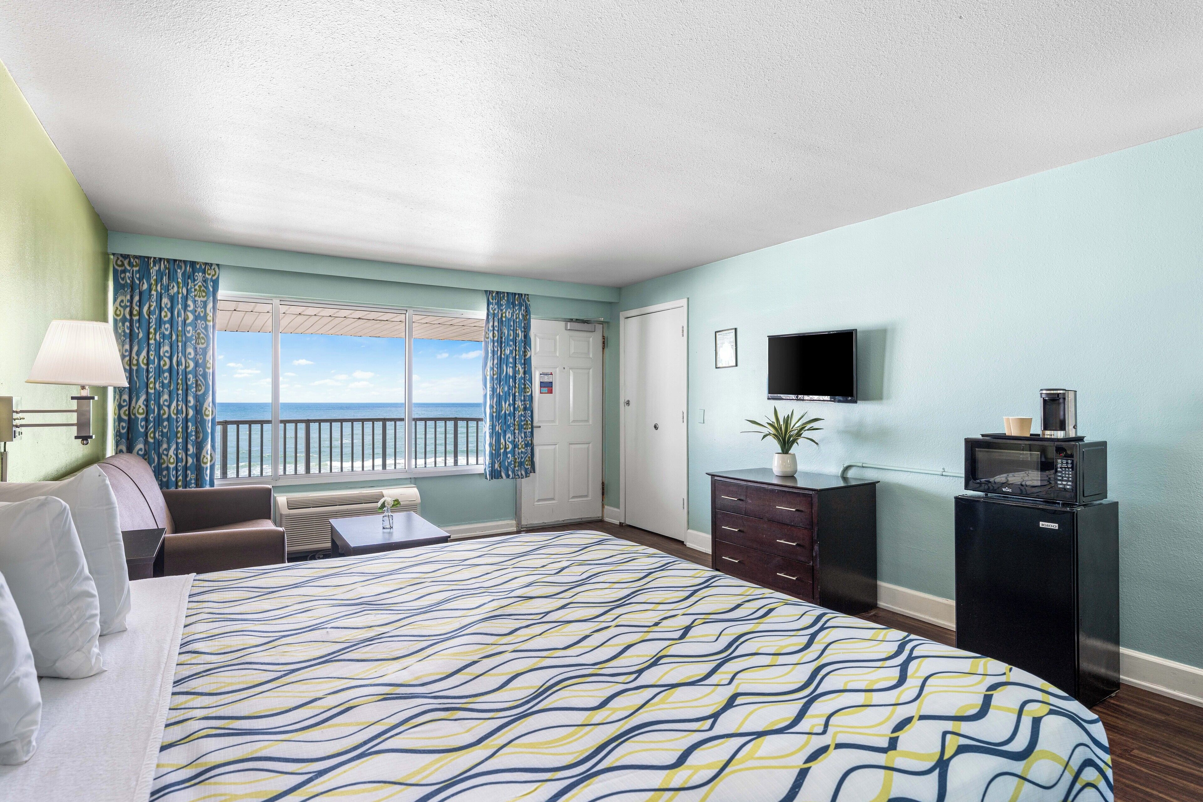 A hotel room with a large bed, a desk, a chair, a television, a microwave, and a view of the ocean.