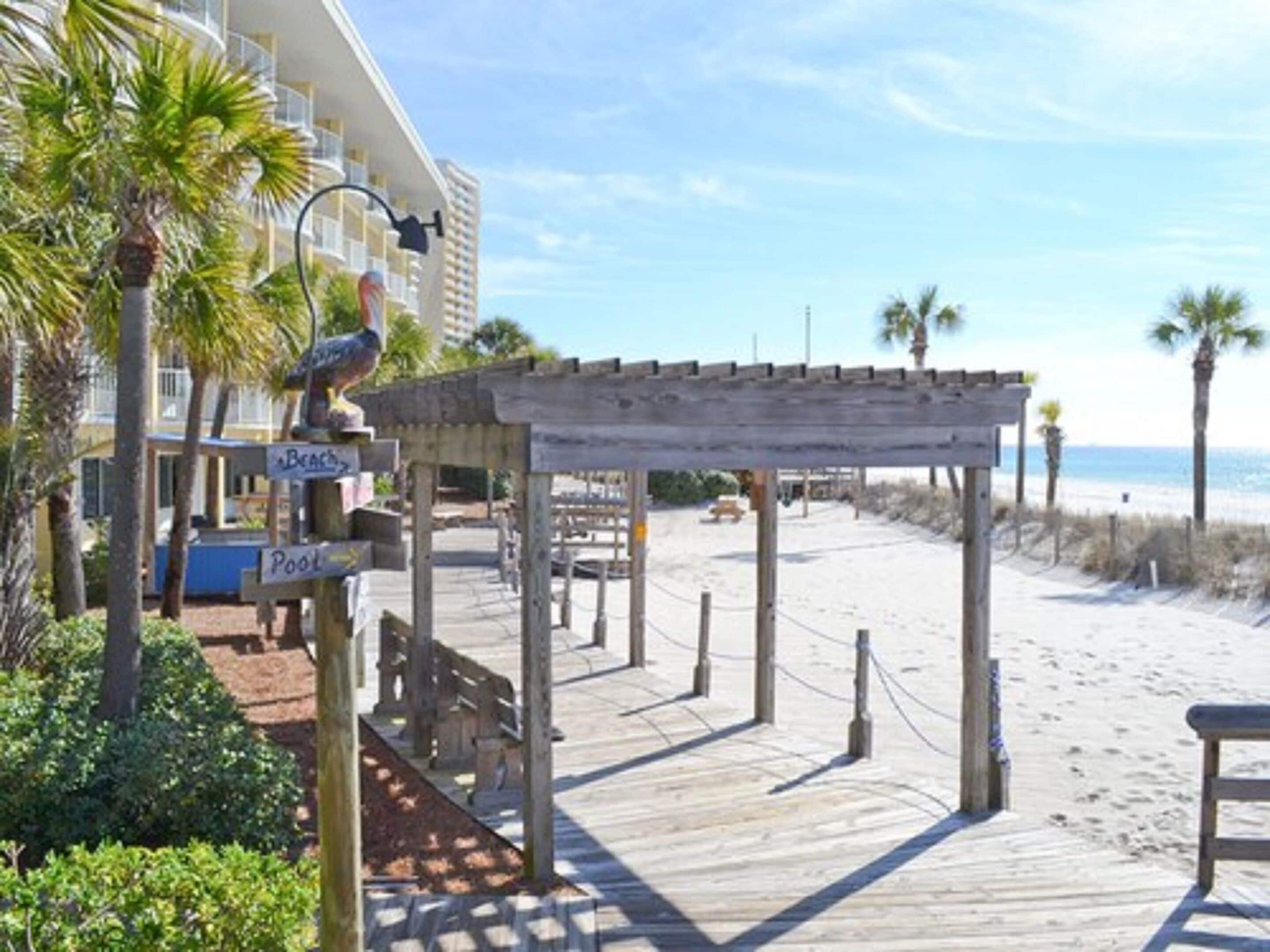 Un paseo marítimo en la playa con una pérgola de madera, palmeras y un letrero que indica una zona de piscina.