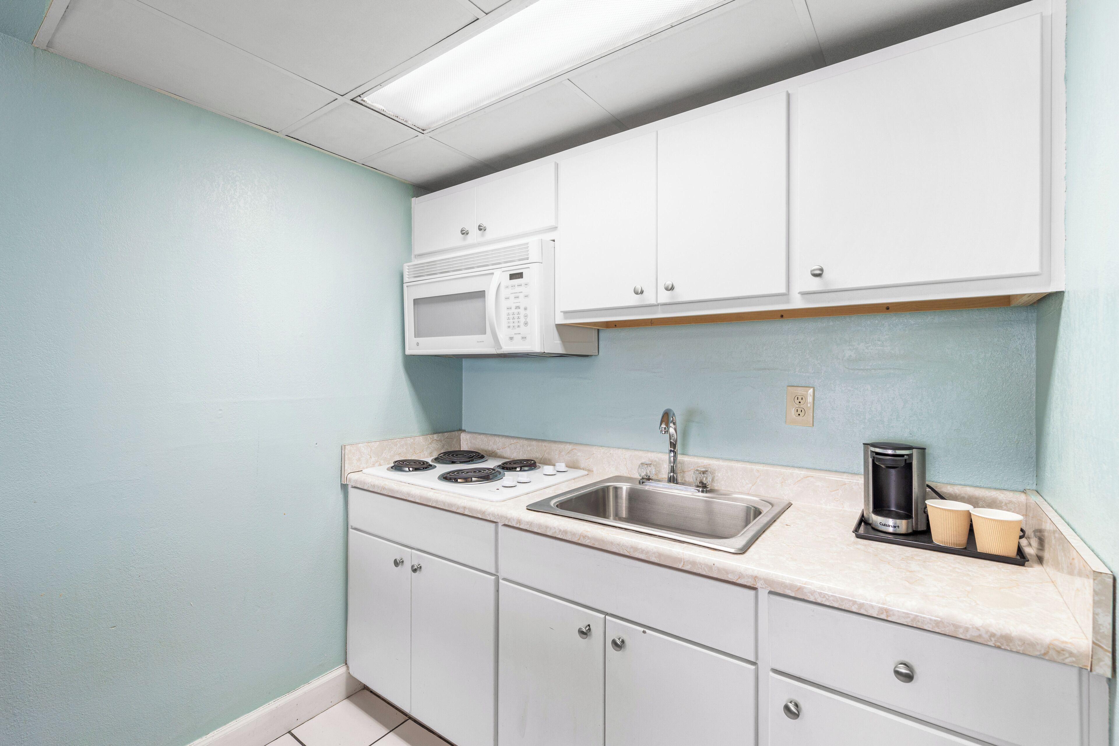 A compact kitchen with white cabinets, a microwave, a stove, a sink, and a coffee maker.