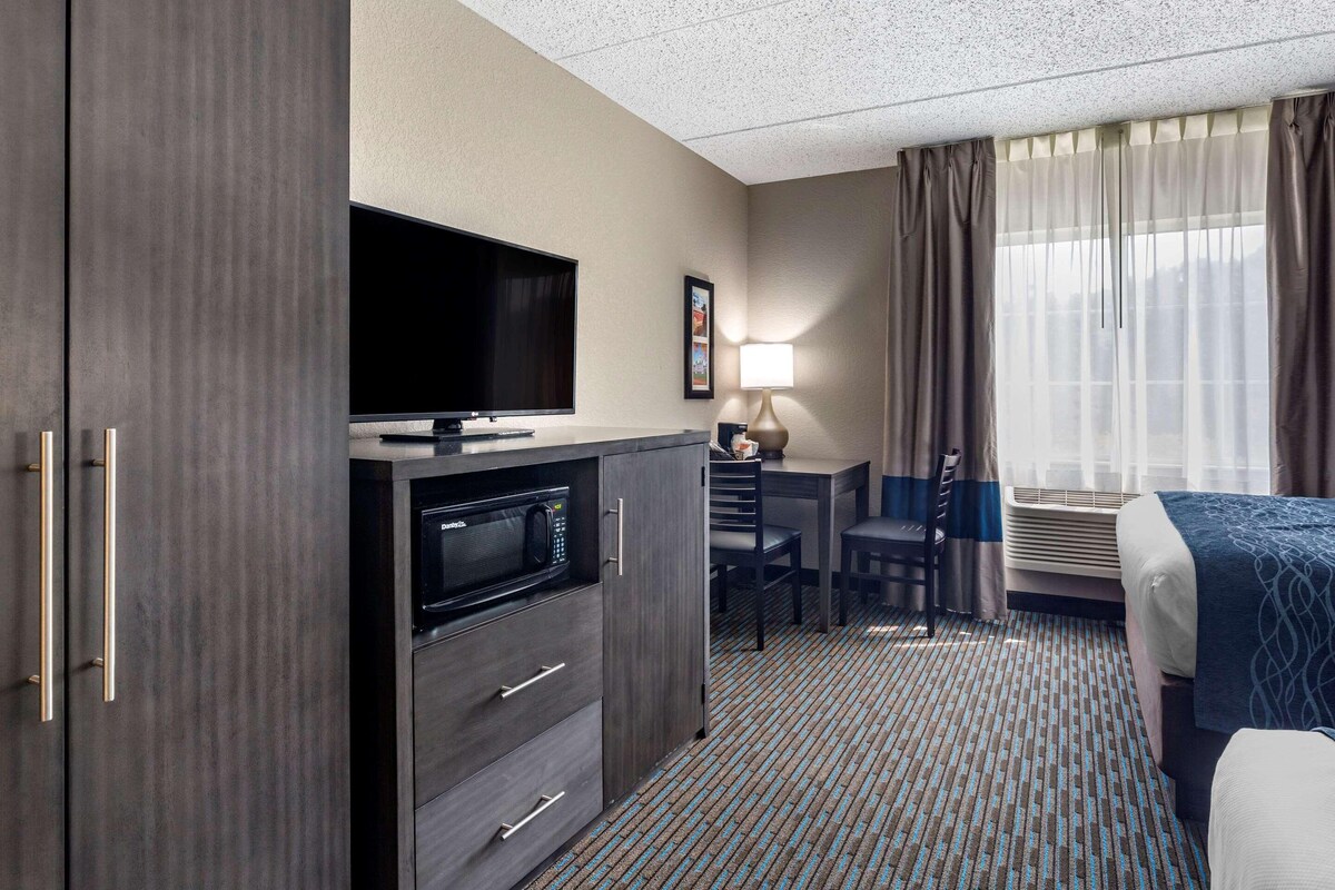 A hotel room with a flat-screen TV, a desk with a lamp, a chair, a bed, and a large window with curtains.