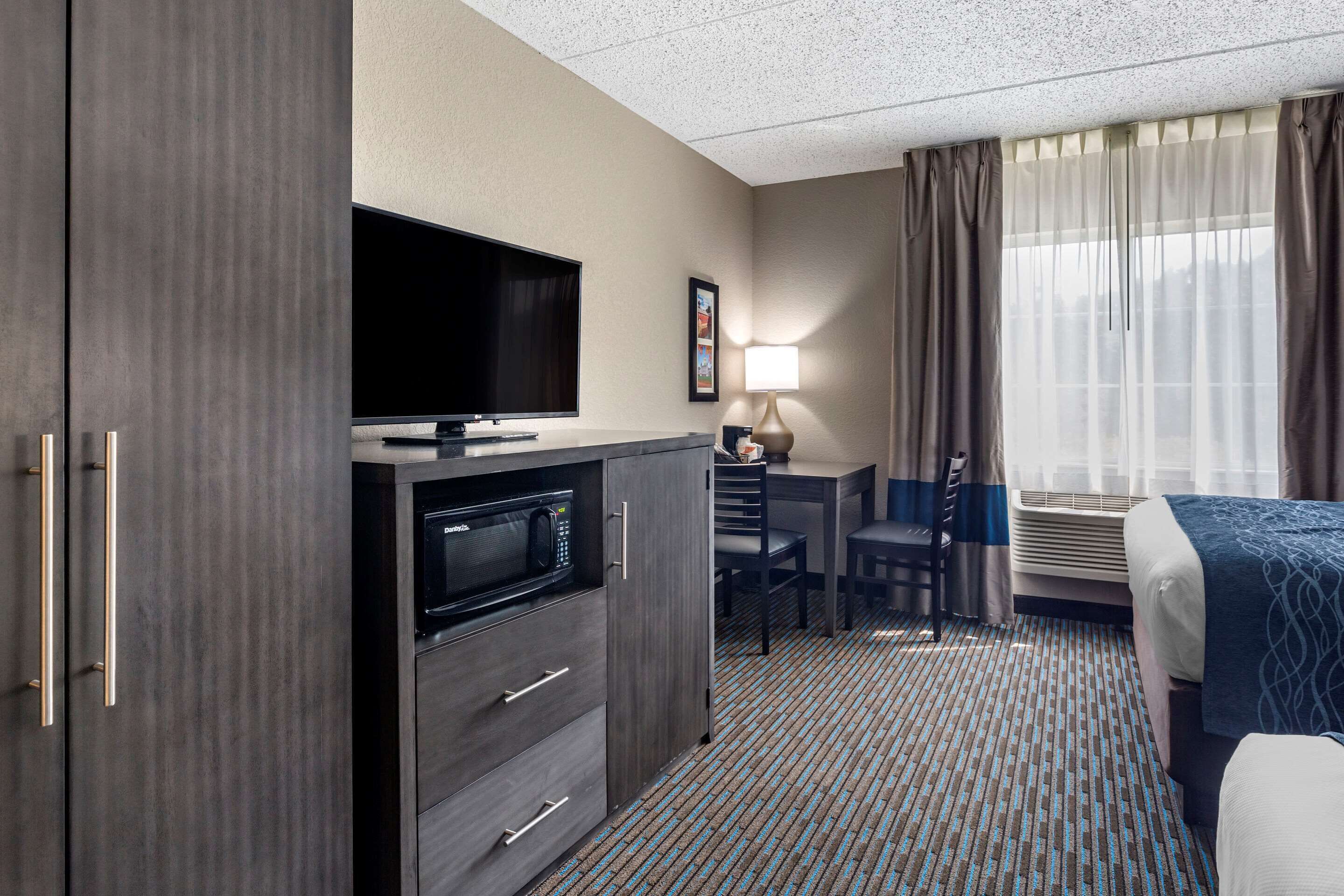 A hotel room with a flat-screen TV, a desk with a lamp, a chair, a bed, and a large window with curtains.