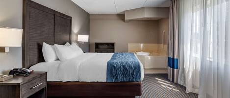 A hotel room with a large bed, a bedside table with a lamp, a small desk with a phone, a TV, and a bathroom with a bathtub and a shower.