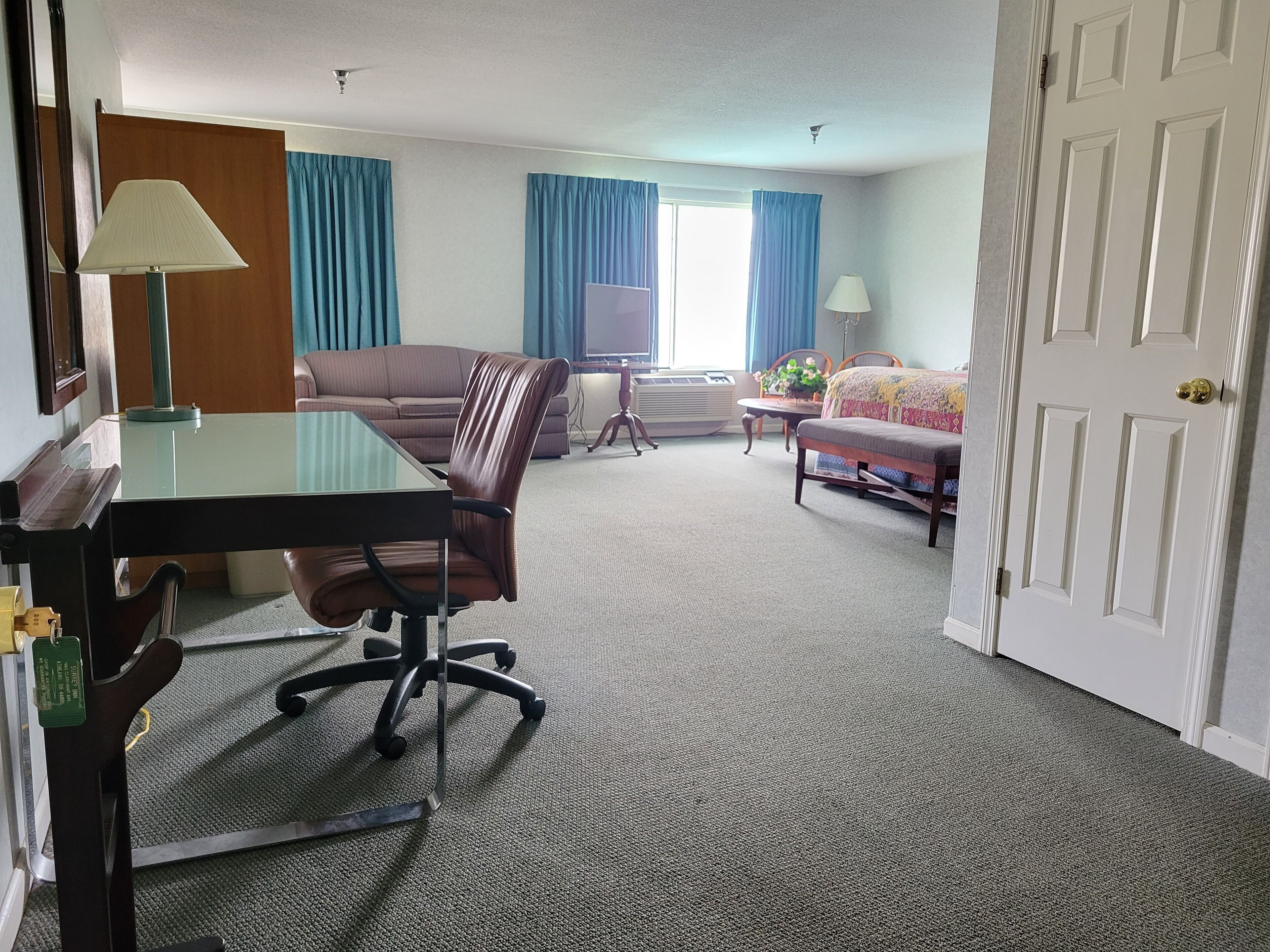 A hotel room with a dining table, a sofa, a chair, a small table with a lamp, and a bed.