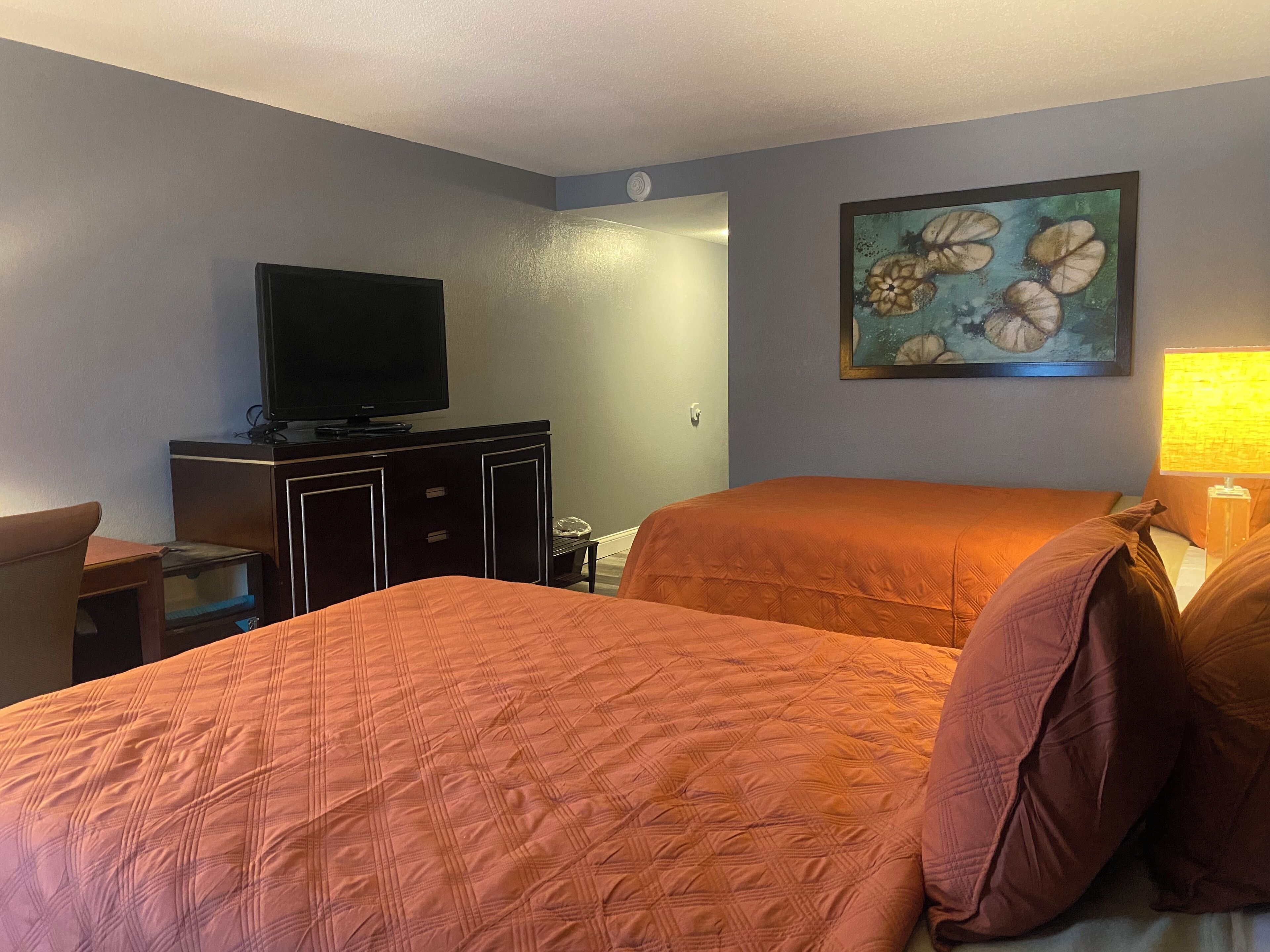 A hotel room with two beds, a television on a dresser, a painting on the wall, and a lamp on the nightstand.