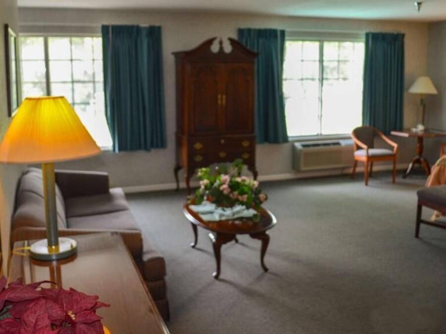 Ein Hotelzimmer mit Sofa, Ohrensessel, einem Holzschrank, einem runden Couchtisch mit Blumen und einem Bett.