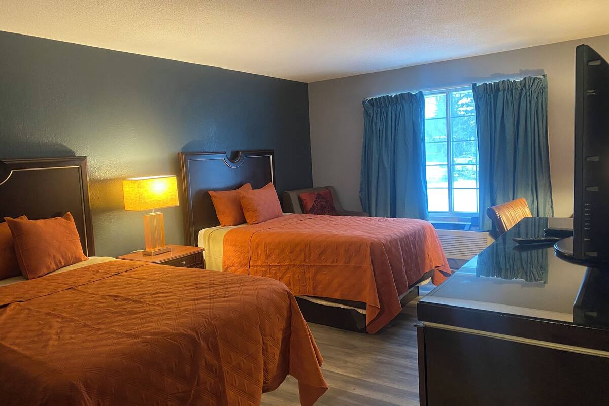 Quarto de hotel com duas camas, uma escrivaninha, uma cadeira, televisão e janela com cortinas.