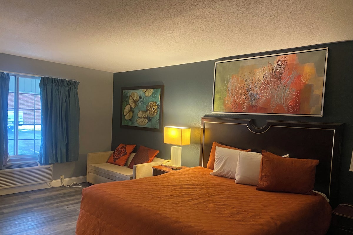 Quarto de hotel com uma cama grande, mesa de cabeceira com abajur, janela com cortinas e um quadro na parede.