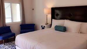 A hotel room with a large bed, two blue armchairs, a nightstand with a lamp, and a window with curtains.