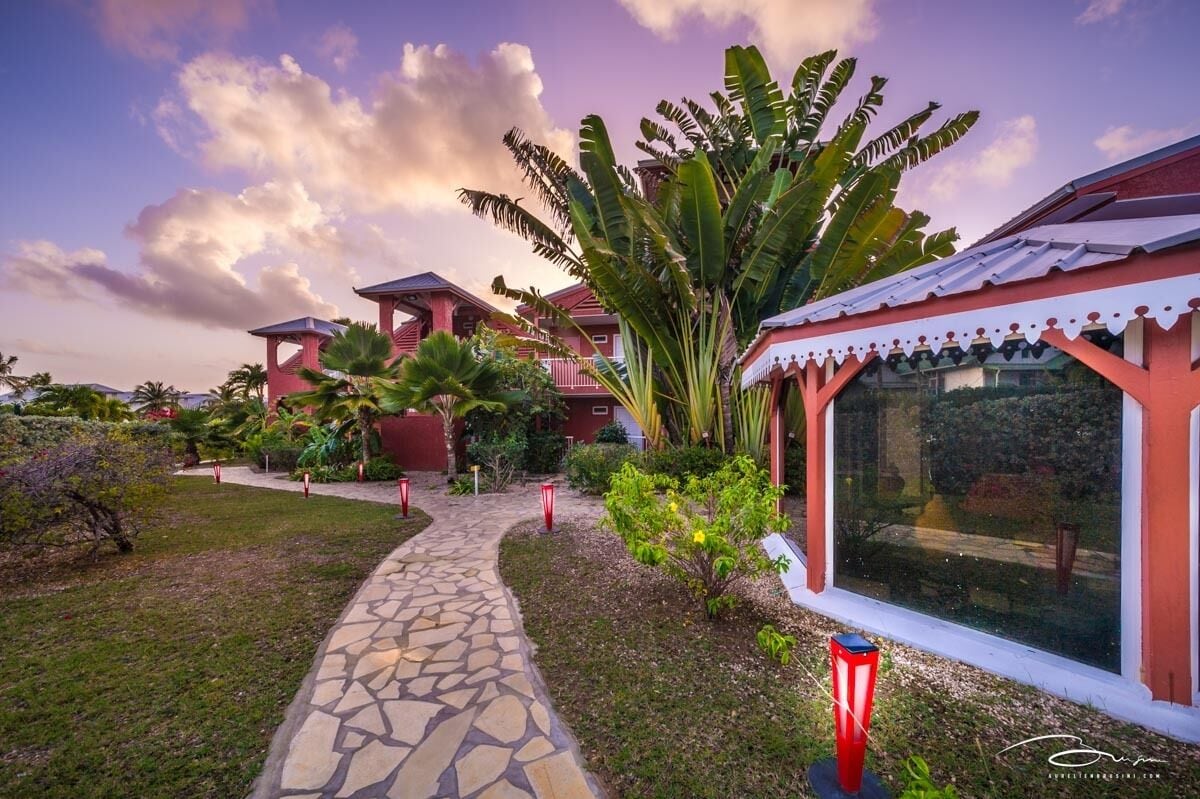 A pathway leading to a building with a red and white facade, surrounded by lush greenery and a clear sky.