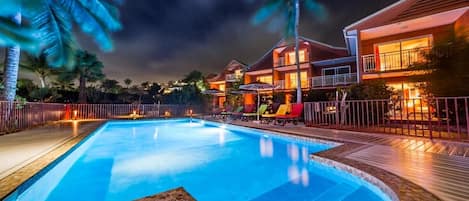 Una piscina con muelle de madera, palmeras y un edificio con varios balcones y zona de estar al aire libre.
