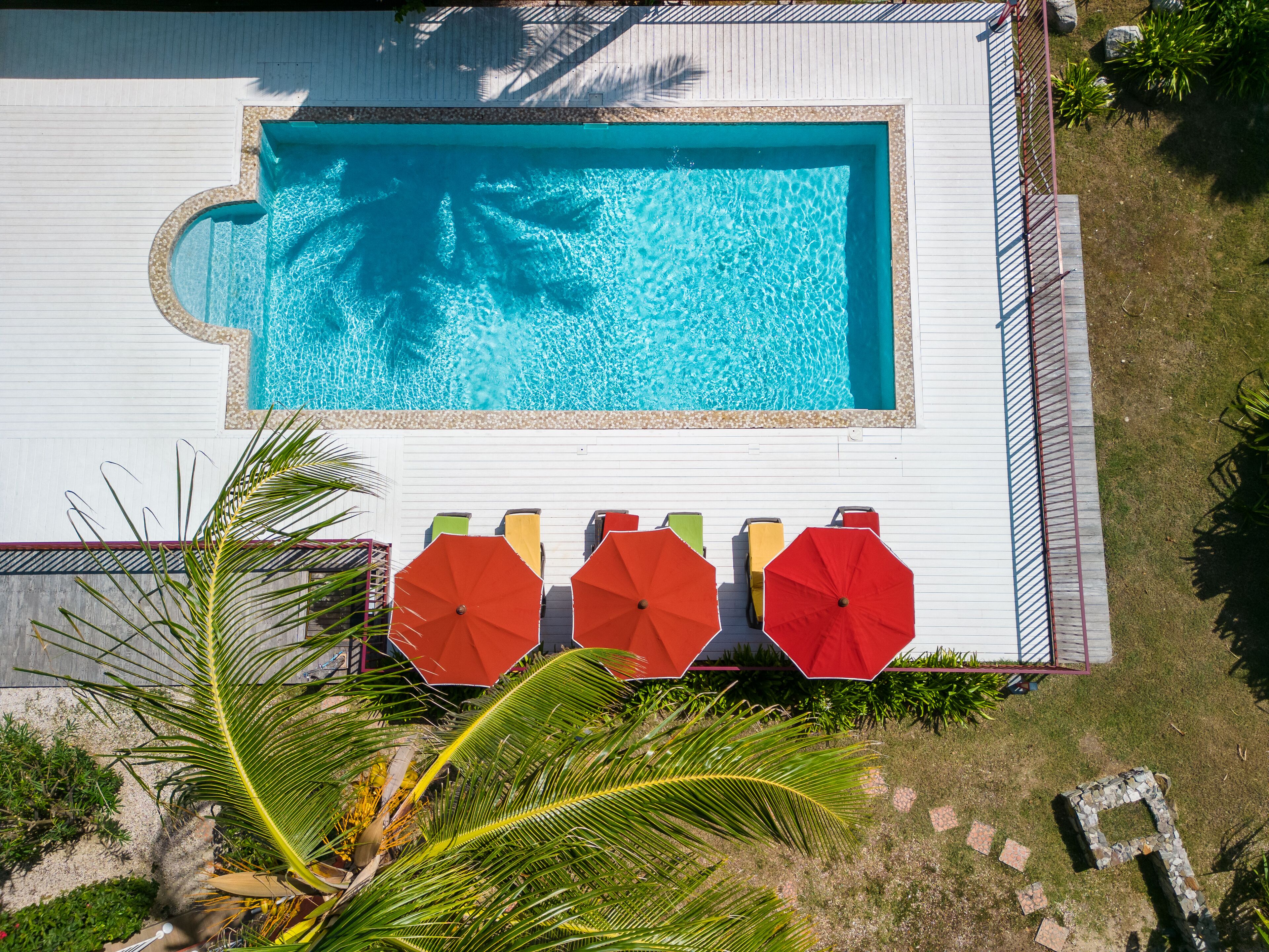 A rectangular swimming pool with a tiled deck, surrounded by a white fence. There are four red umbrellas and a few lounge chairs along the deck.