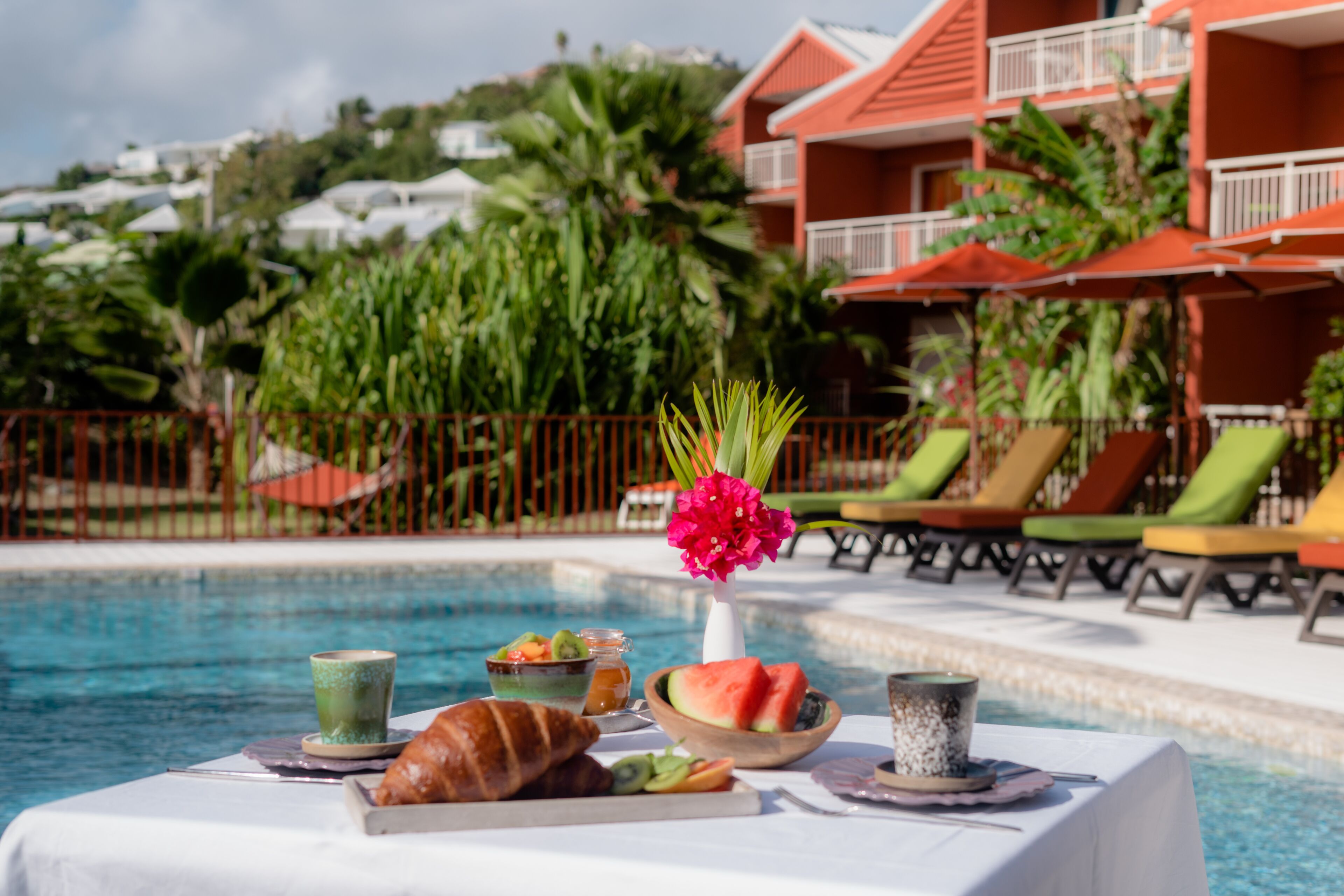 A poolside breakfast setup with a croissant, fruit bowl, and drinks on a table.