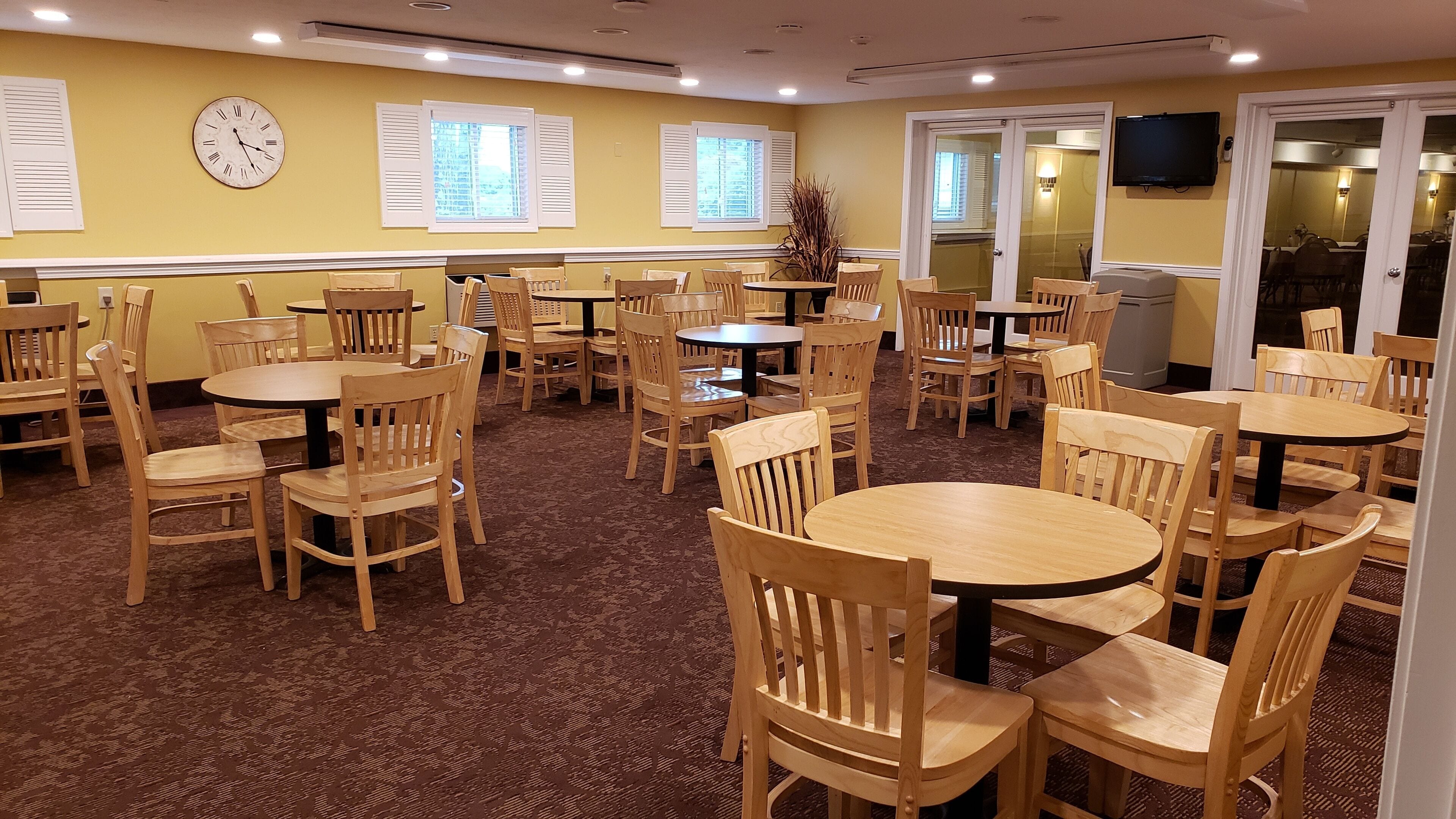 Un espace repas avec plusieurs tables rondes et des chaises en bois, une horloge au mur et un téléviseur fixé au mur.