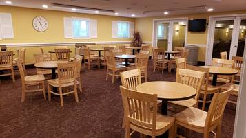 Un espace repas avec plusieurs tables rondes et des chaises en bois, une horloge au mur et un téléviseur fixé au mur.