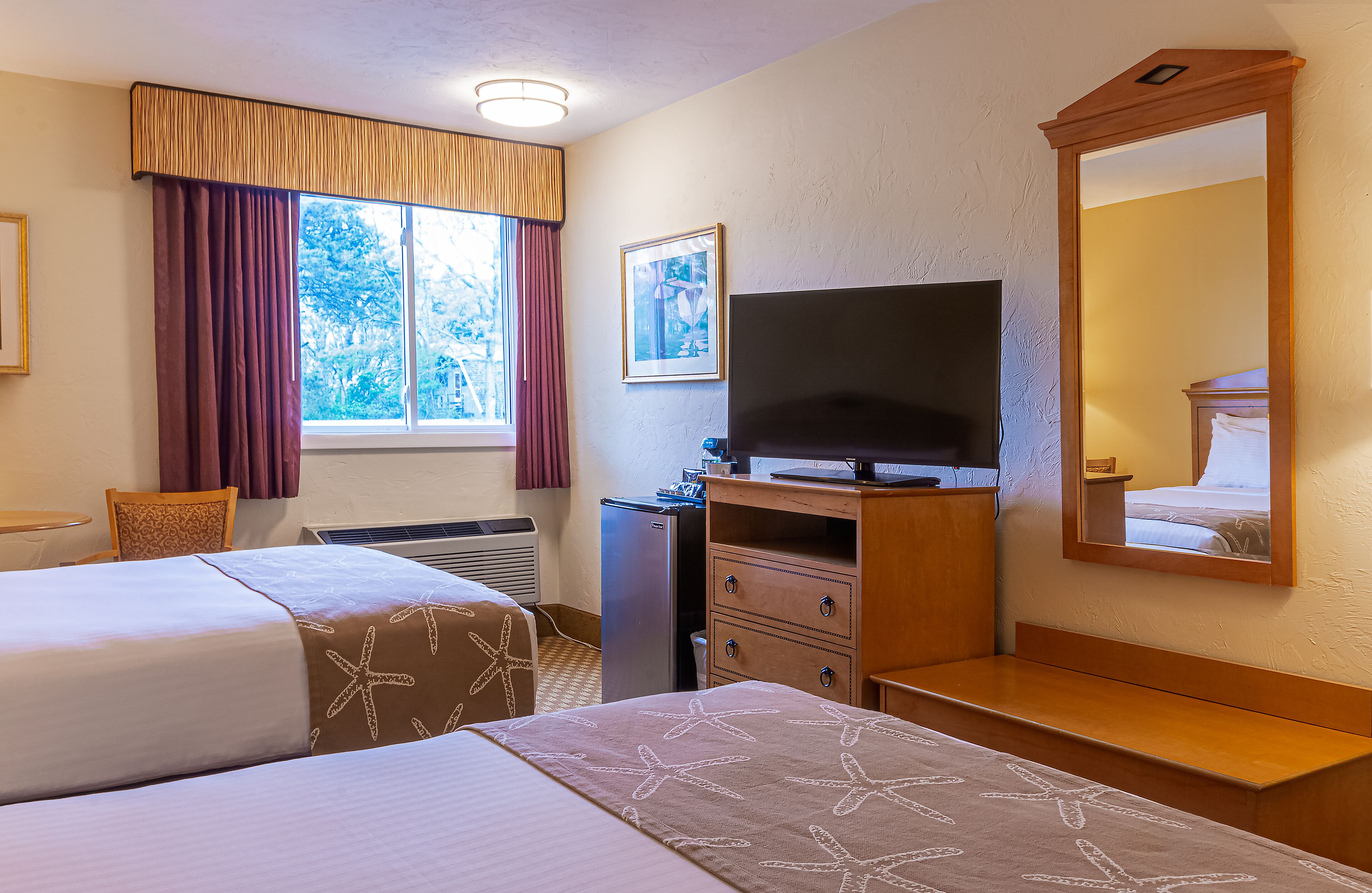 A hotel room with two beds, a television, a dresser, a mirror, and a window with curtains.