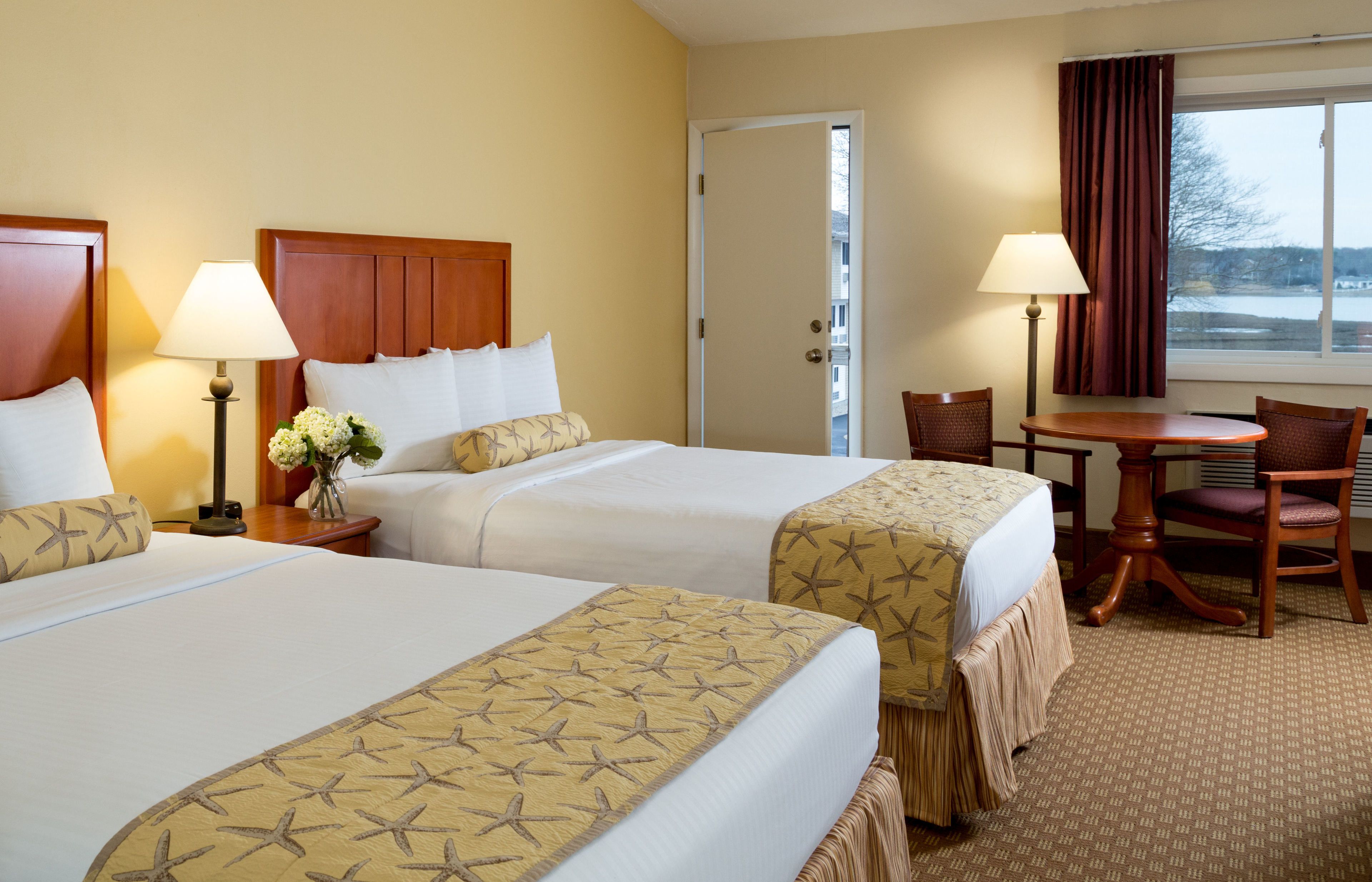 Quarto de hotel com duas camas, uma escrivaninha, uma cadeira e uma janela com cortinas.