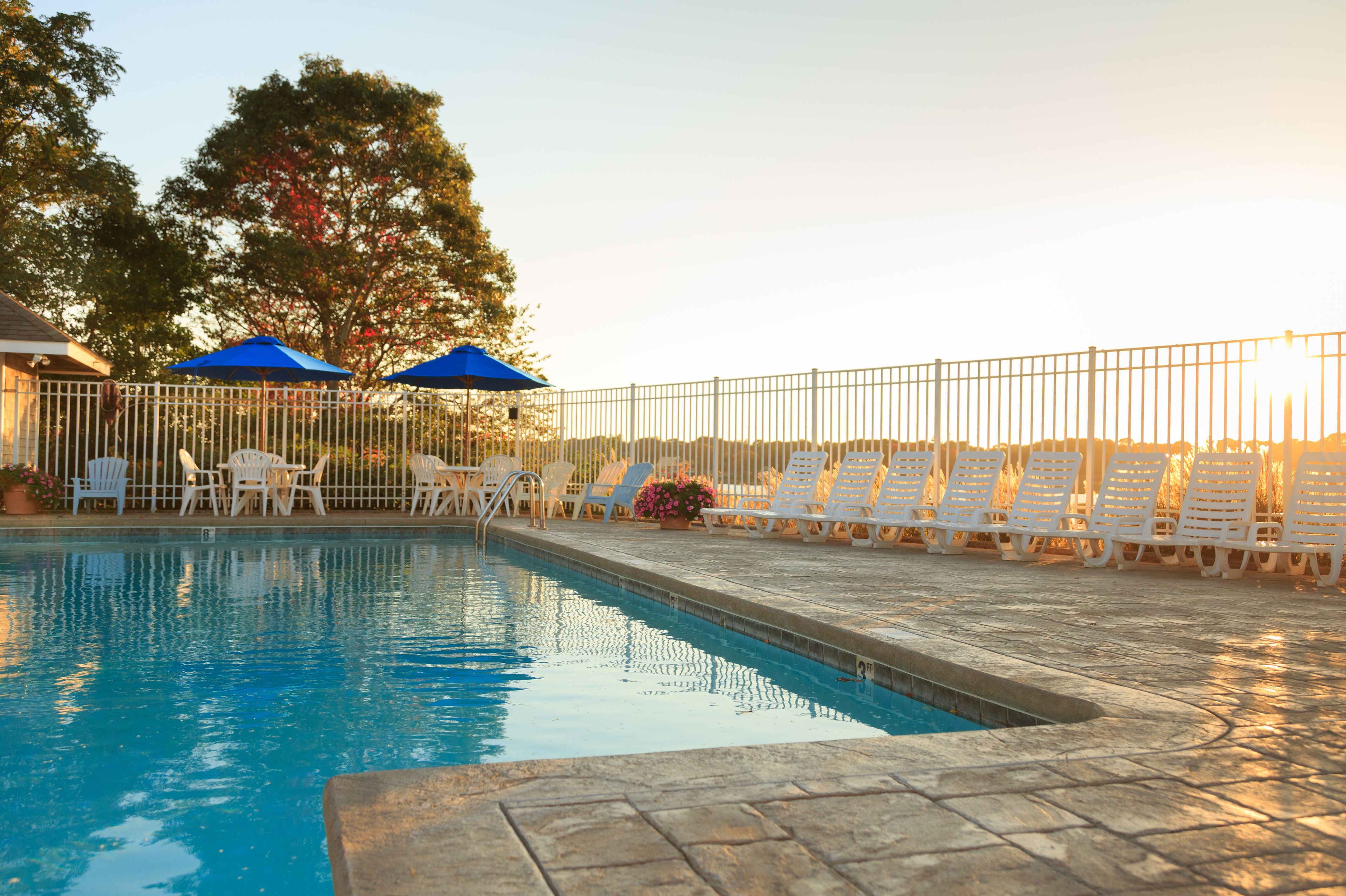 A swimming pool with white lounge chairs and blue umbrellas, surrounded by a metal fence.