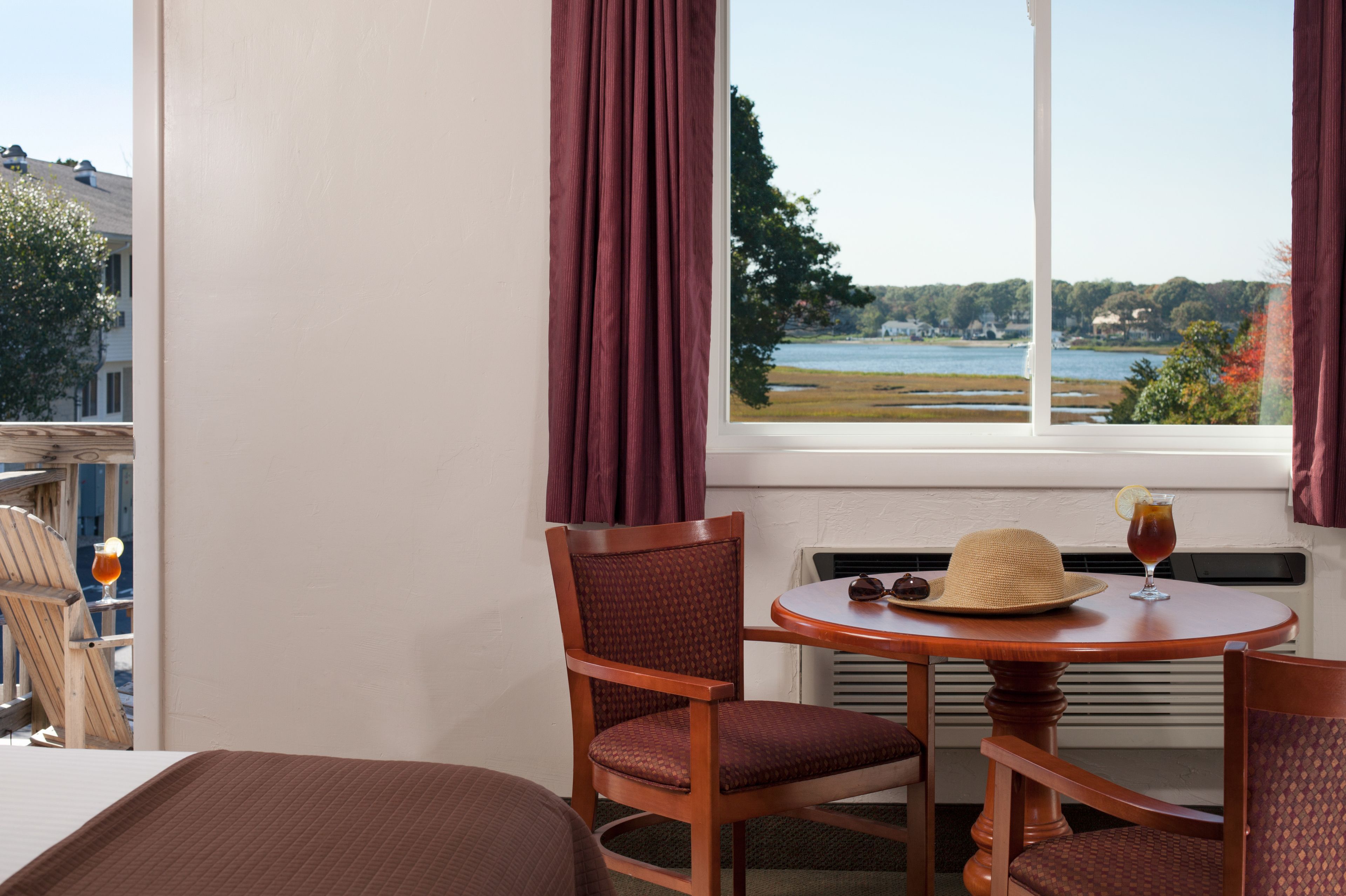 A hotel room with a view of a lake, a round wooden table with a hat and a drink, and two wooden chairs.