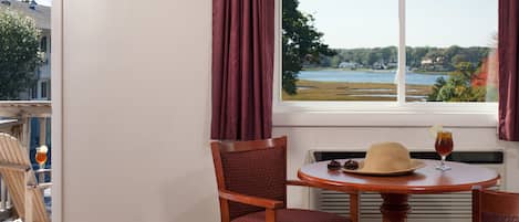 A hotel room with a view of a lake, a round wooden table with a hat and a drink, and two wooden chairs.