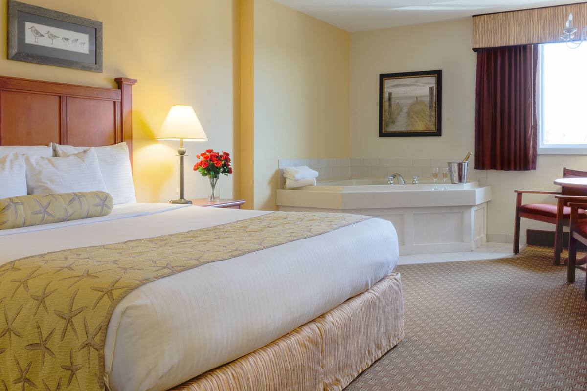 Hotelkamer met een groot bed, een zithoek met een fauteuil, een bureau, een lamp, een vaas met bloemen, een schilderij, een badkuip en een raam met gordijnen.