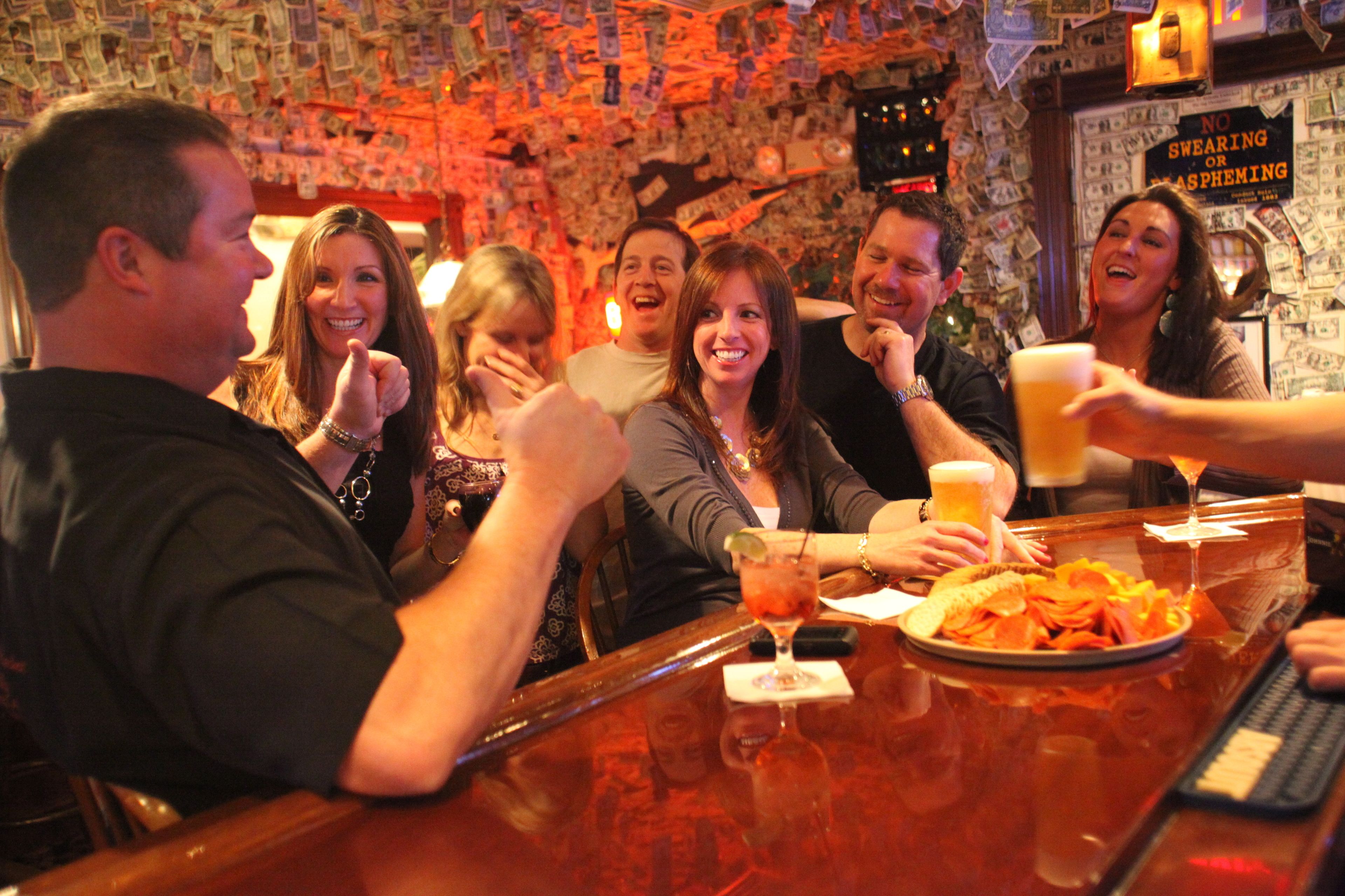 A group of people at a bar with a wall covered in dollar bills.
