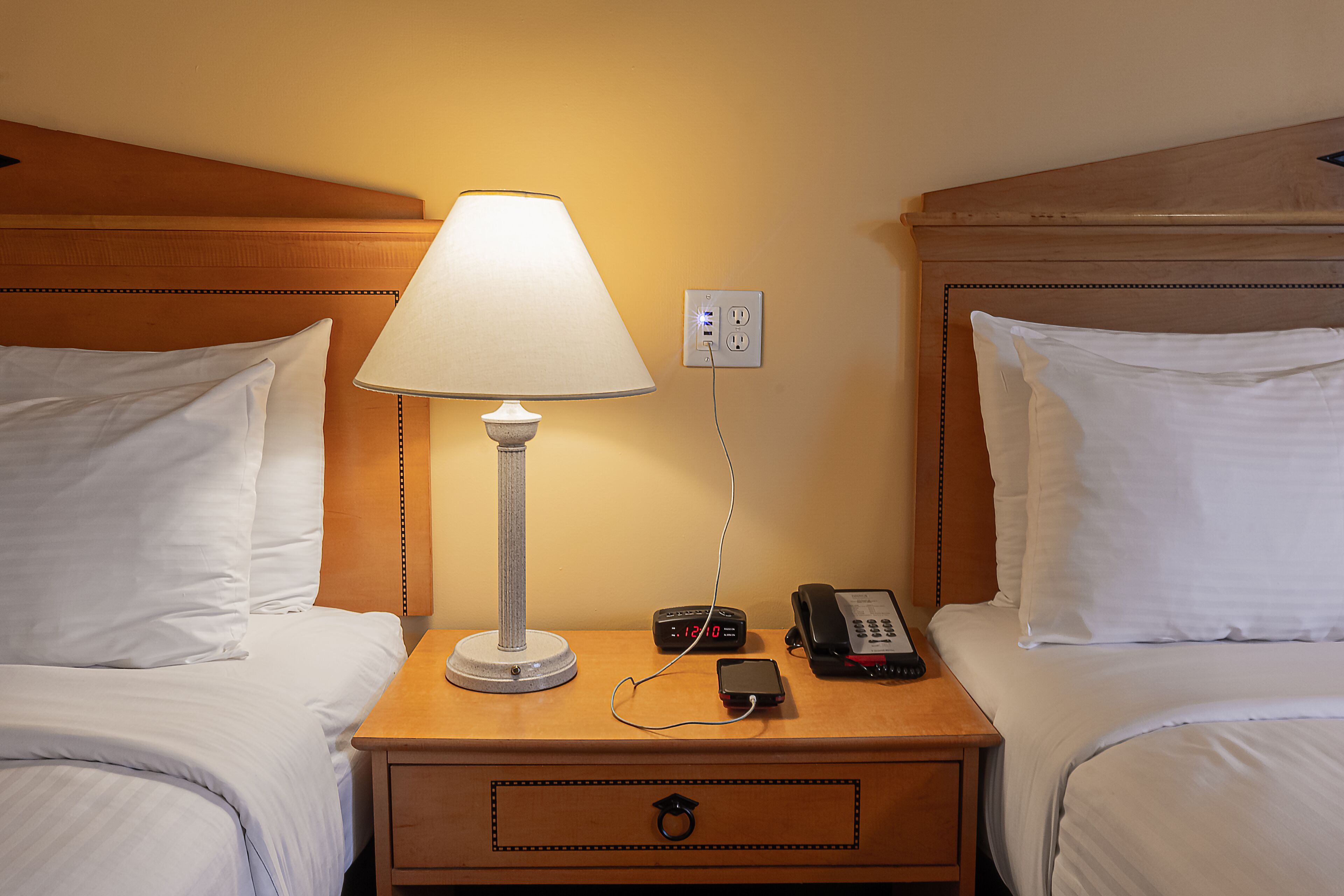 Quarto de hotel com duas camas, uma mesa de cabeceira com abajur, telefone, relógio e tomada.