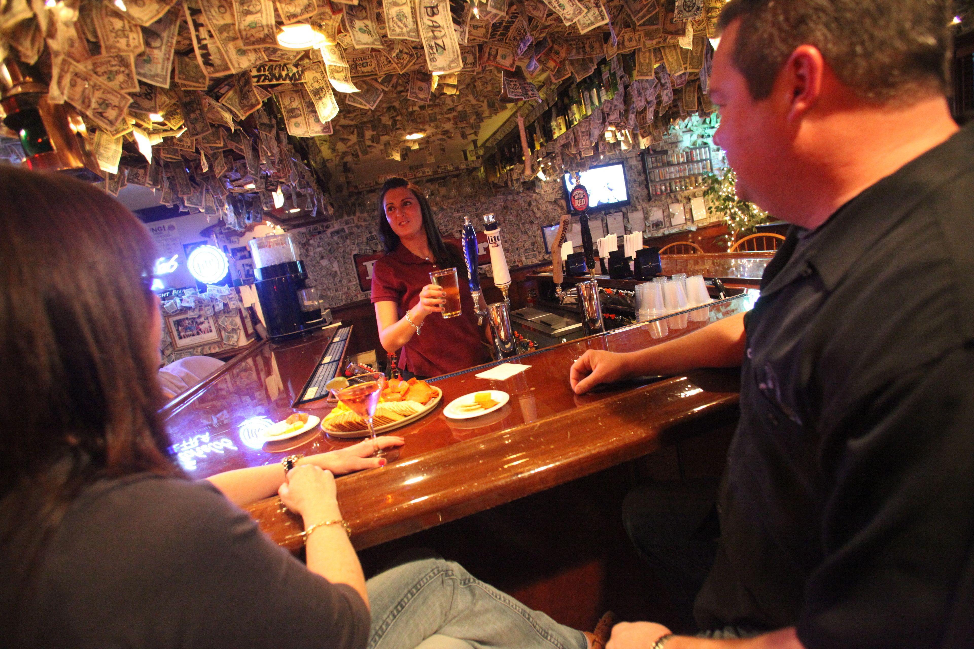 A bar with a ceiling covered in dollar bills, a bartender, and patrons seated at the bar.