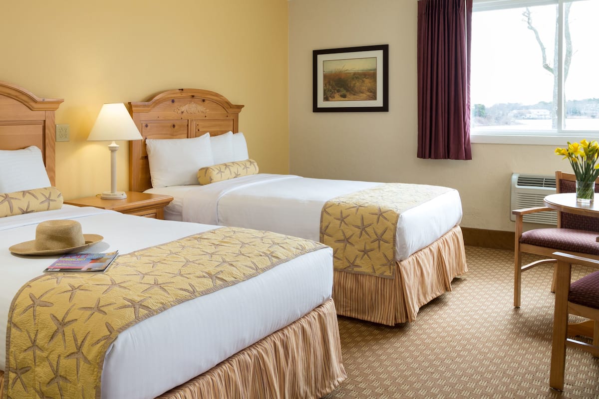 Een hotelkamer met twee bedden, een nachtkastje met een lamp, een stoel, een tafel met een bloemetjesvaas, en een raam met uitzicht.