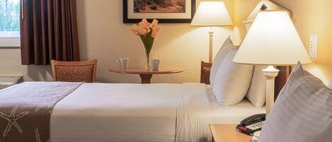 A hotel room with a bed, a bedside table with a lamp, a vase with flowers, and a framed picture on the wall.