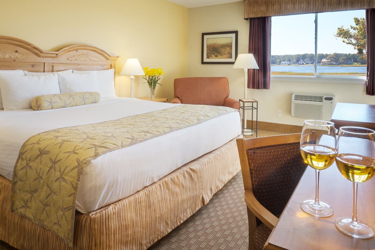 Een hotelkamer met een bed, een stoel, een tafel met twee glazen wijn, een lamp en een raam met uitzicht op een meer.